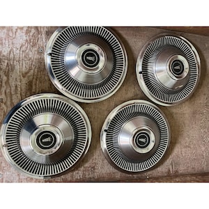 Puede incluir: Cuatro tapacubos cromados vintage con un diseño radial y un emblema central. Los tapacubos están dispuestos sobre una superficie de madera, mostrando su calidad reflectante y estética clásica. Los tapacubos son probablemente para un coche.