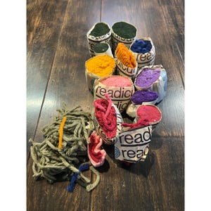 Könnte beinhalten: Eine Sammlung bunter Garnbündel und kleiner Papierverpackungen, gefüllt mit verschiedenen Garnfarben. Auf den Verpackungen steht das Wort "read". Die Garnfarben umfassen Grün, Gelb, Orange, Rosa, Lila, Blau und Rot.