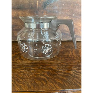 May include: A clear glass coffee carafe with a gray handle and a silver band. White floral designs decorate the carafe. Measurement markings are visible on the side, with a capacity of up to 10 cups. The carafe is designed for brewing coffee.
