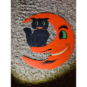 May include: A vintage Halloween decoration featuring a black cat with green eyes perched on an orange crescent moon. The moon has a smiling face with a green eye and black eyelashes. The cat has a mischievous grin. A festive, retro-style decoration.