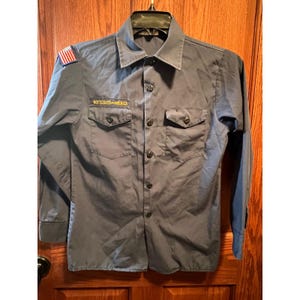May include: A long-sleeved, button-down shirt in a solid slate blue color. The shirt features a pointed collar, two front chest pockets with buttoned flaps, and a patch on the right shoulder with the American flag. A patch reads BOYSCOUTS OF AMERICA.