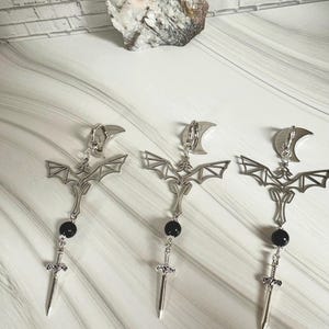 Könnte beinhalten: Silberfarbene Ohrringe mit einem Fledermaus-Design, einem Halbmond, einer schwarzen Perle und einem Schwertanhänger. Die Ohrringe werden auf einer weiß-grauen Oberfläche präsentiert, mit einem dekorativen Stein im Hintergrund.