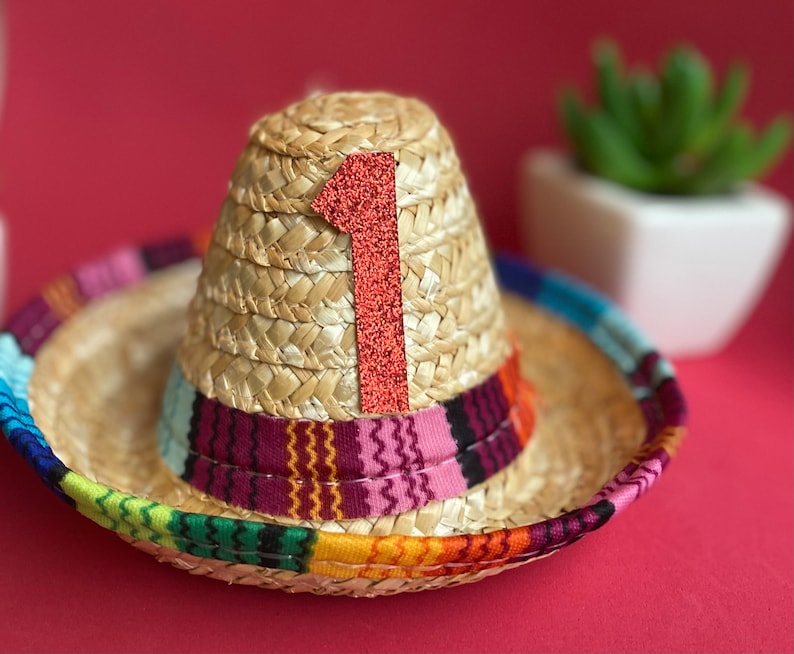 Pu&ograve; includere: Un piccolo sombrero di paglia con una fascia colorata e un numero uno in feltro rosso sulla parte superiore.