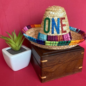 Pu&ograve; includere: Un piccolo sombrero di paglia con una fascia a righe colorate e la parola "ONE" in lettere glitterate. Il cappello &egrave; su una scatola di legno.