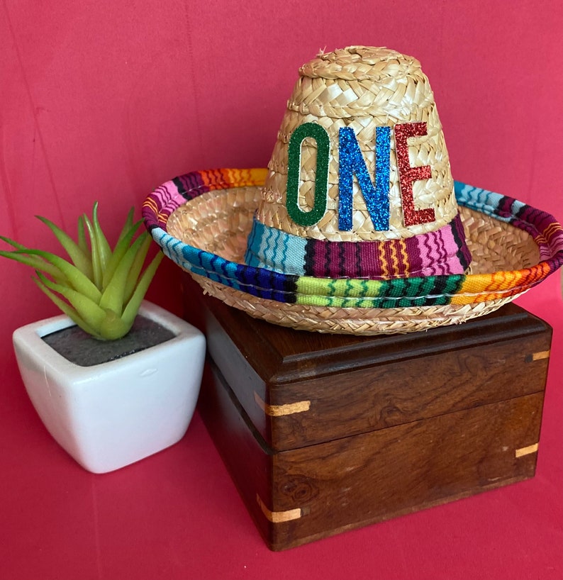 Pu&ograve; includere: Un piccolo sombrero di paglia con una fascia a righe colorate e la parola "ONE" in lettere glitterate. Il cappello &egrave; su una scatola di legno.