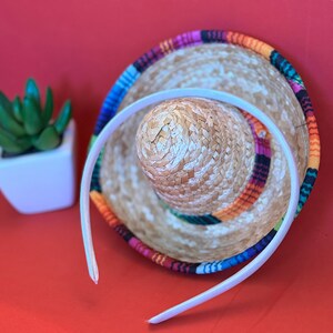 Pu&ograve; includere: Un cerchietto bianco con un sombrero colorato sotto. Il sombrero ha una base di paglia intrecciata e una fascia di tessuto colorata.
