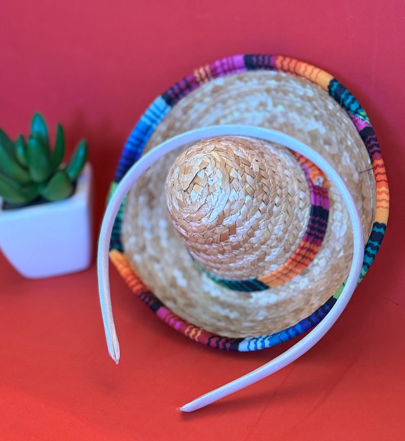 Pu&ograve; includere: Un cerchietto bianco con un sombrero colorato sotto. Il sombrero ha una base di paglia intrecciata e una fascia di tessuto colorata.