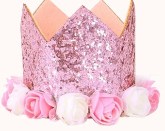 Può includere: Corona rosa glitter con un bordo di fiori bianchi e rosa. La corona è fatta di cartone e ha una punta appuntita.