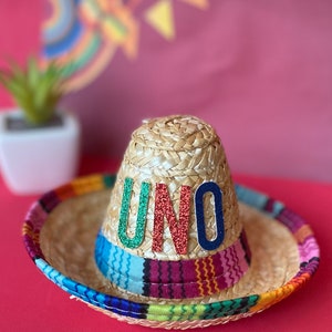 Pu&ograve; includere: Un sombrero di paglia con una fascia a righe colorate e la parola "UNO" in lettere glitter sulla corona.