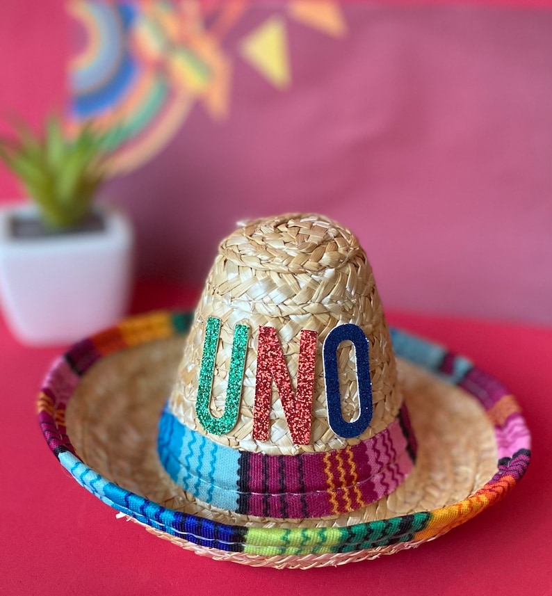 Pu&ograve; includere: Un sombrero di paglia con una fascia a righe colorate e la parola "UNO" in lettere glitter sulla corona.