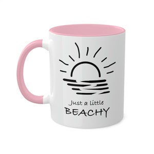 May include: White ceramic mug with a pink interior and handle. The mug features a black graphic of a sun over water and the text "Just a little BEACHY".