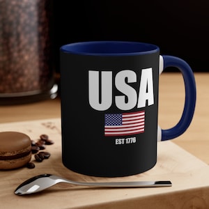 May include: Black coffee mug with a blue rim and handle. The mug has a white graphic of the word "USA" with an American flag and the text "EST 1776" underneath.