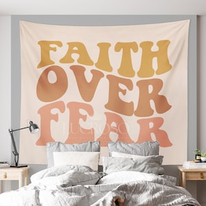 May include: A tapestry with the text "FAITH OVER FEAR" in a retro font. The letters are in shades of orange, yellow, and pink. The tapestry is hanging on a wall behind a bed with gray bedding.