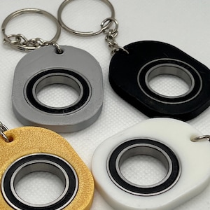 May include: Four keychains with a bearing design. The keychains are in gold, gray, black, and white. Each keychain has a silver metal ring and chain attached to the top. The bearing is black and silver.