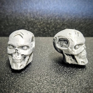 Puede incluir: Dos figuras de calaveras grises impresas en 3D con rasgos faciales detallados, que incluyen cuencas de los ojos, una cavidad nasal y dientes. Una calavera está erguida y la otra está inclinada hacia un lado. Las calaveras están sobre una superficie oscura y texturizada.