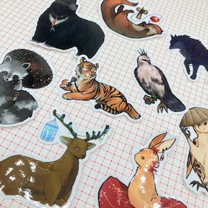 May include: A collection of whimsical animal stickers, including a tiger, raccoon, deer, and bear. The stickers feature detailed illustrations with vibrant colors and are arranged on a red and white checkered background.