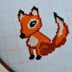 Peut inclure: Une broderie au point de croix représentant un renard assis, la queue enroulée autour de son corps. Le renard est orange avec des accents blancs et des yeux noirs. La broderie est réalisée sur un tissu blanc à motif quadrillé.