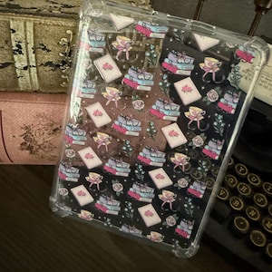 May include: Clear phone case with a repeating pattern of books, teacups, and cards in pink, blue, and yellow. The case has a black and white background. The case is made of clear plastic.
