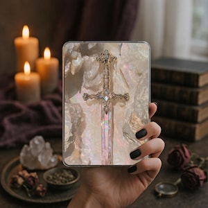 May include: A decorative phone case featuring a detailed sword design. The sword has a gold-colored hilt and a pink, iridescent blade. The background has a pearlescent, marbled effect. The case is held in a hand with black nail polish.