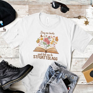 May include: White t-shirt with a brown graphic of an open book with flowers and butterflies. The text "Buy me books and tell me to STFUATTDLAGG" is printed on the shirt.