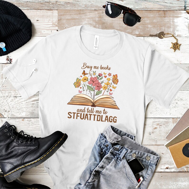 May include: White t-shirt with a brown graphic of an open book with flowers and butterflies. The text "Buy me books and tell me to STFUATTDLAGG" is printed on the shirt.