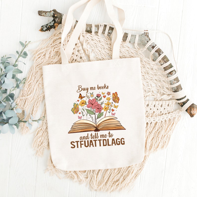 May include: A white tote bag with a brown print of an open book with flowers coming out of it. The text on the bag reads "Buy me books and tell me to STFUATTDLAGG".
