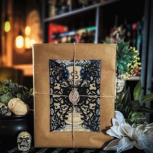 May include: A wrapped package with a decorative black and blue design, tied with twine. The package has a vintage aesthetic, with a wax seal and a floral design. The background includes a black cauldron, skulls, and white flowers.