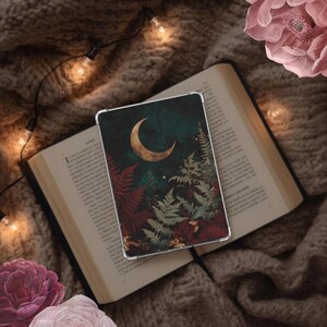May include: A clear tablet case featuring a dark green and black design with a gold crescent moon, ferns, and stars. The case rests on an open book with a soft, brown knitted blanket in the background. Fairy lights and pink flowers add to the aesthetic.