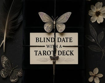 May include: A black velvet box tied with black string, adorned with a metal butterfly and a dagger charm. A card reads "BLIND DATE WITH A TAROT DECK." The image includes black feathers, dried flowers, and a butterfly.