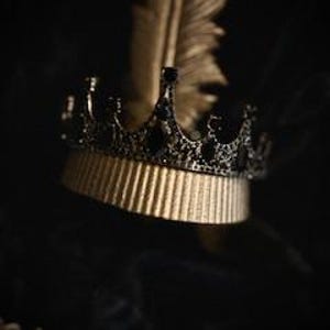 Op de afbeelding: Een zwarte kroon met zwarte edelstenen en een gouden basis. De kroon ligt op een zwarte veer.