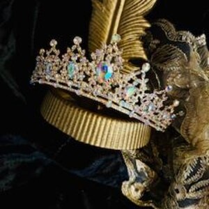 Op de afbeelding: Een gouden kroon met heldere, iriserende stenen en een rij kleinere stenen. De kroon staat op een gouden standaard.