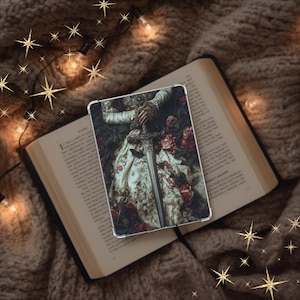 May include: A clear tablet case featuring a detailed image of a person in a floral dress holding a sword, set against a backdrop of red roses. The case rests on an open book with a brown cover, surrounded by a cozy, textured blanket and decorative star-shaped lights.