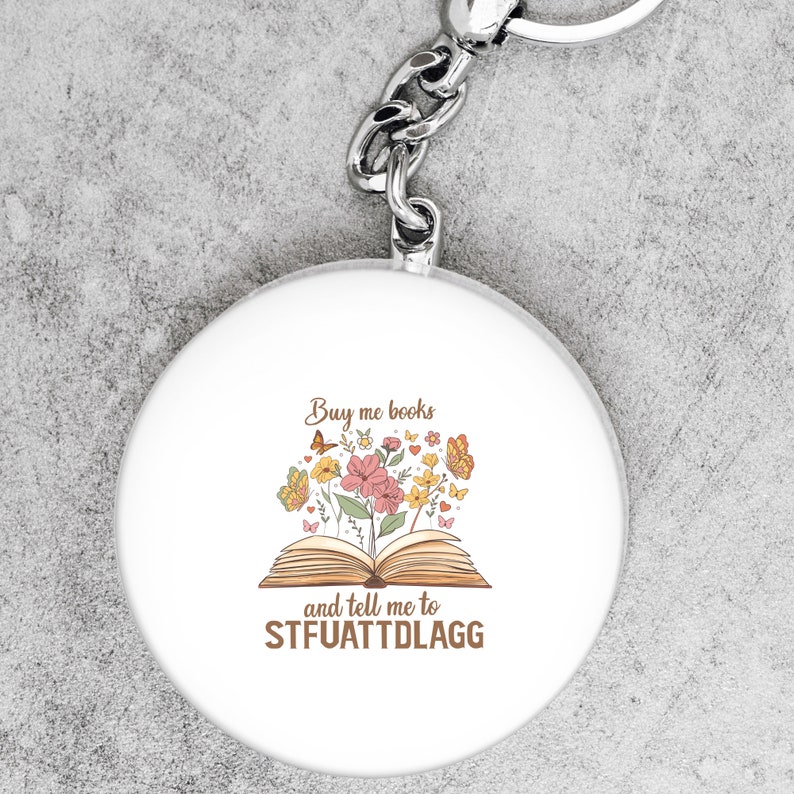 May include: White round keychain with a book and flowers design. The text reads "Buy me books and tell me to STFUATTDLGG".