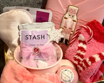 May include: A pink gift box filled with self-care items. Includes a pink eye mask, tea bags, a pink bow, a pink mug, a bath bomb, a pink and white striped candy cane, a pink and red sock, a pink bow, a snowflake, and a pink nutcracker.
