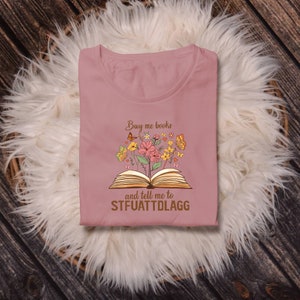 May include: A pink t-shirt with a graphic of an open book with flowers and butterflies. The text on the shirt reads "Buy me books and tell me to STFUATTDLAGG".