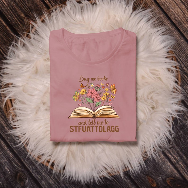 May include: A pink t-shirt with a graphic of an open book with flowers and butterflies. The text on the shirt reads "Buy me books and tell me to STFUATTDLAGG".
