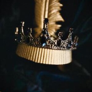 Op de afbeelding: Een gouden kroon met zwarte accenten en blauwe edelstenen. De kroon ligt op een gouden oppervlak met een gouden veer op de achtergrond.