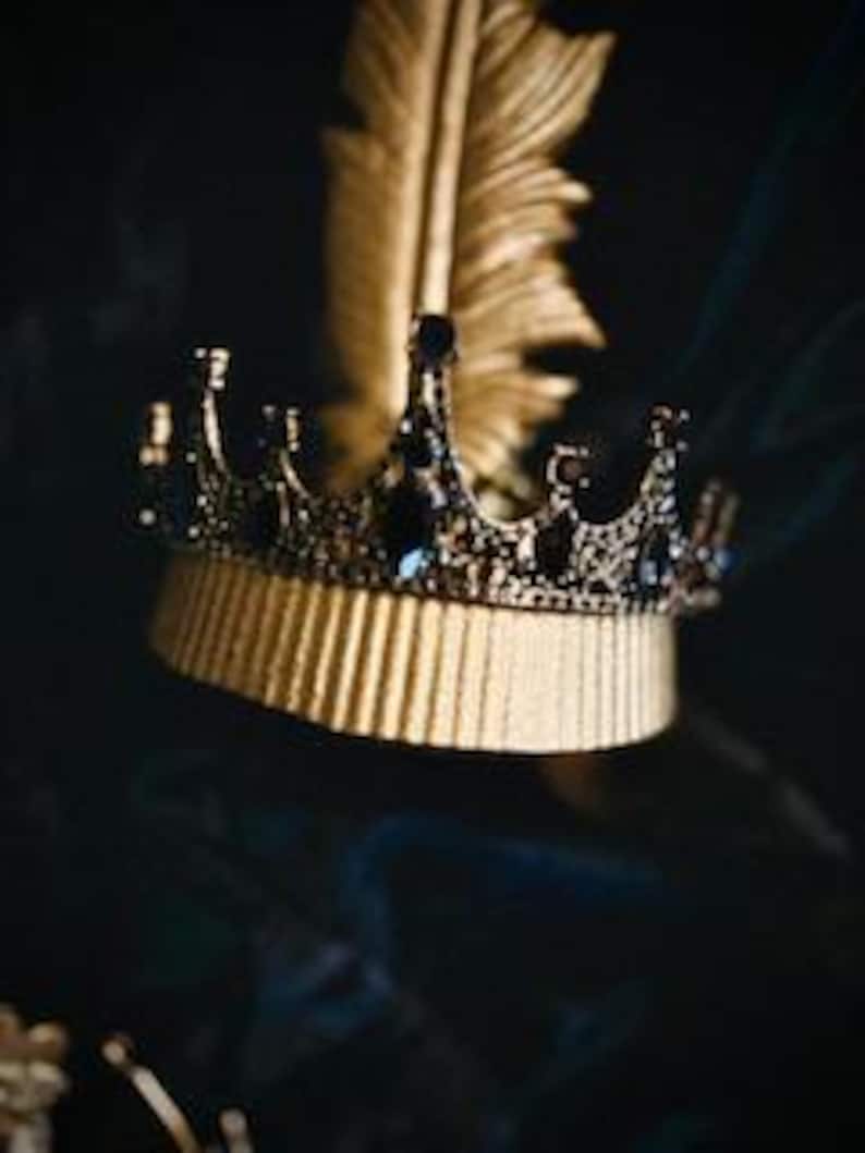 Op de afbeelding: Een gouden kroon met zwarte accenten en blauwe edelstenen. De kroon ligt op een gouden oppervlak met een gouden veer op de achtergrond.