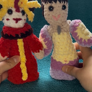 May include: Two handmade crochet dolls. One doll is dressed in red with yellow accents and a yellow and brown headpiece. The other doll is dressed in yellow and purple with a brown head and arms. Both dolls have black button eyes and are holding hands.