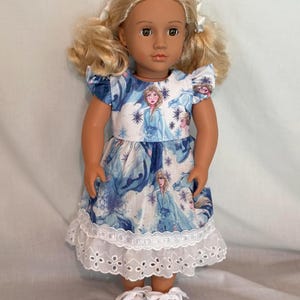 Puede incluir: Mu&ntilde;eca con coletas rubias, vestida con un vestido azul y blanco con un dise&ntilde;o de princesa y un dobladillo de encaje blanco. La mu&ntilde;eca lleva zapatillas blancas. El vestido presenta un dise&ntilde;o de princesa.