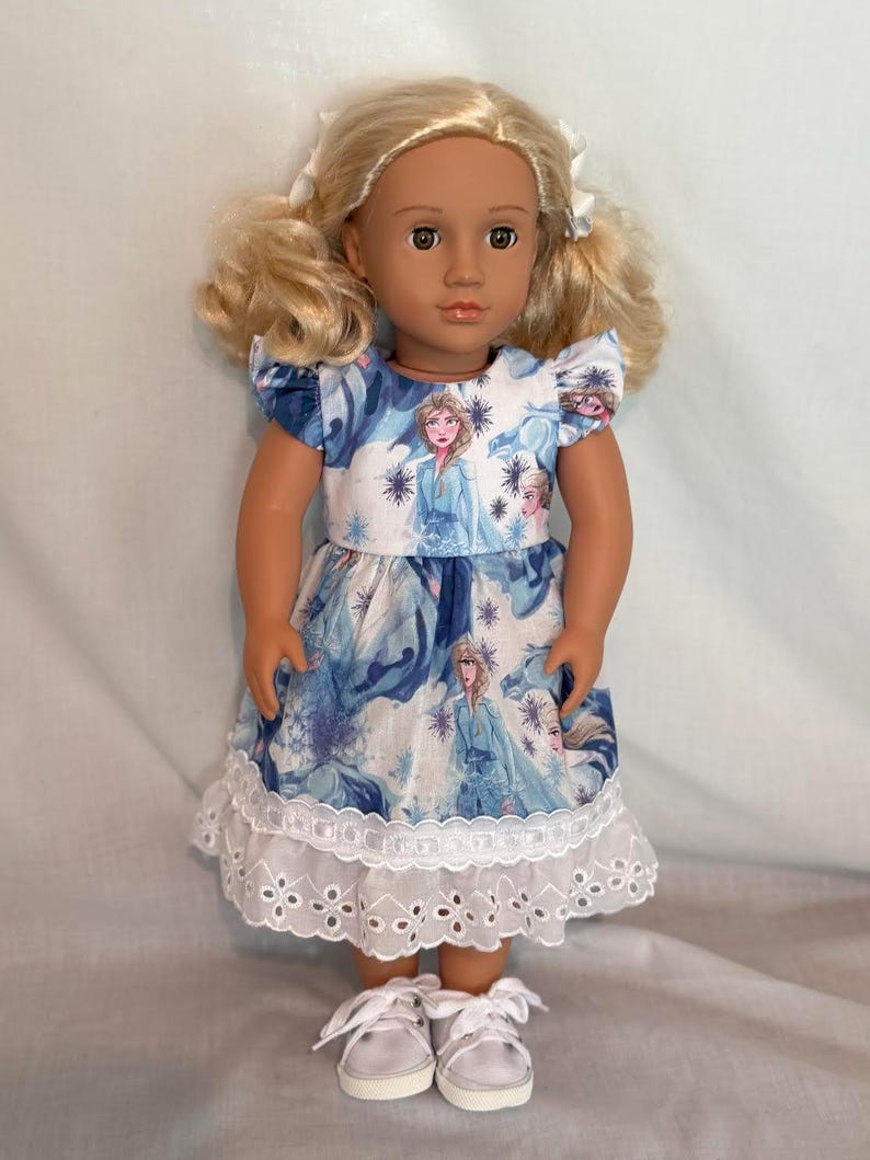 Puede incluir: Mu&ntilde;eca con coletas rubias, vestida con un vestido azul y blanco con un dise&ntilde;o de princesa y un dobladillo de encaje blanco. La mu&ntilde;eca lleva zapatillas blancas. El vestido presenta un dise&ntilde;o de princesa.