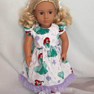 May include: A doll with blonde hair styled in pigtails, wearing a white dress with a print of Ariel from The Little Mermaid. The dress has a green bodice and sleeves, a purple lace hem, and white sneakers.