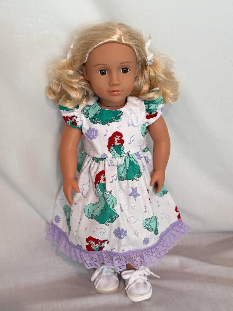 May include: A doll with blonde hair styled in pigtails, wearing a white dress with a print of Ariel from The Little Mermaid. The dress has a green bodice and sleeves, a purple lace hem, and white sneakers.