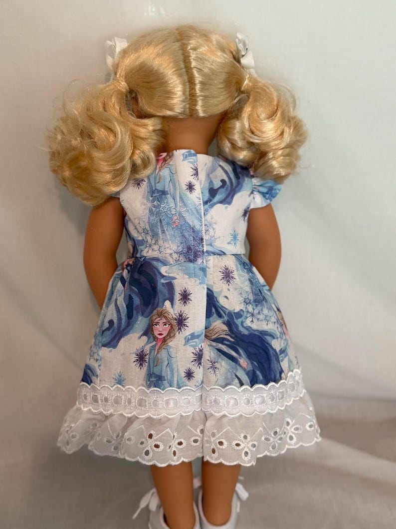 Puede incluir: Una mu&ntilde;eca con un vestido blanco con un estampado de dibujos animados azul y blanco. El vestido tiene mangas cortas y un ribete de encaje calado blanco en el dobladillo. La mu&ntilde;eca tiene el pelo rubio peinado con coletas.