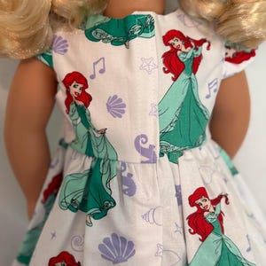 May include: A white doll dress featuring a repeating pattern of a red-haired princess in a green dress, musical notes, seashells, and stars. The dress has a green trim and is designed for a doll.