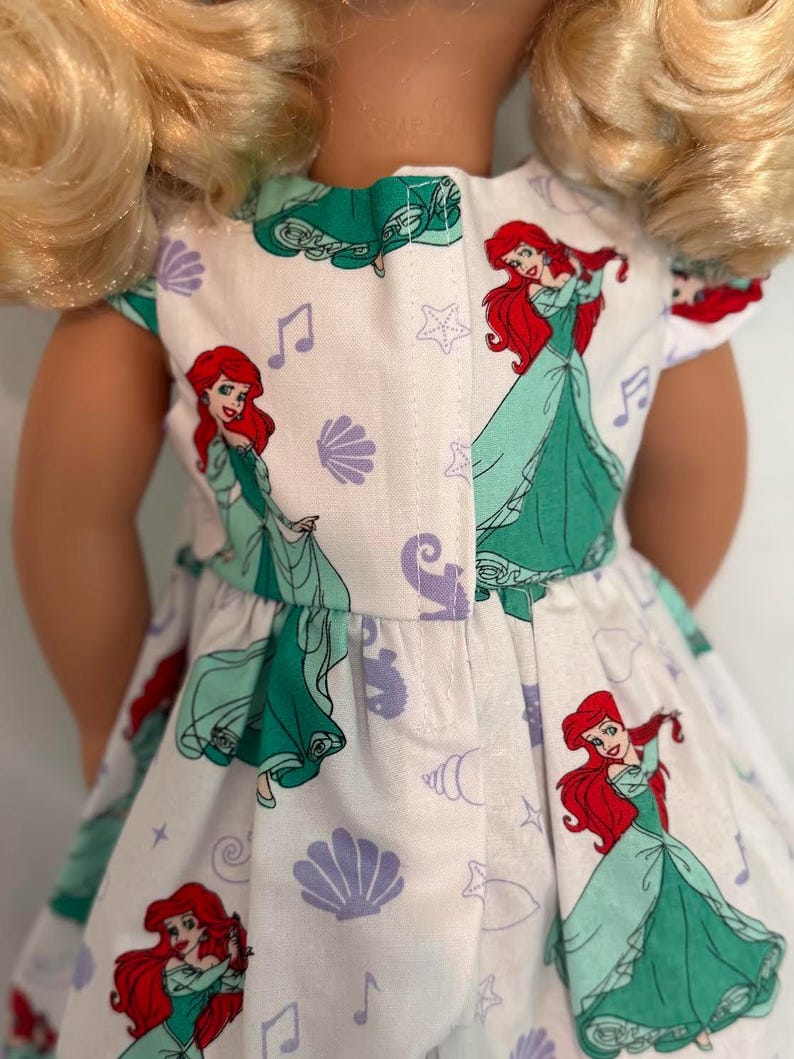 May include: A white doll dress featuring a repeating pattern of a red-haired princess in a green dress, musical notes, seashells, and stars. The dress has a green trim and is designed for a doll.