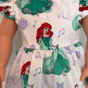 May include: A white doll dress featuring a repeating pattern of a red-haired princess in a green dress, musical notes, castles, seashells, and starfish. The dress has short sleeves and a gathered waist. The doll's blonde hair is visible.