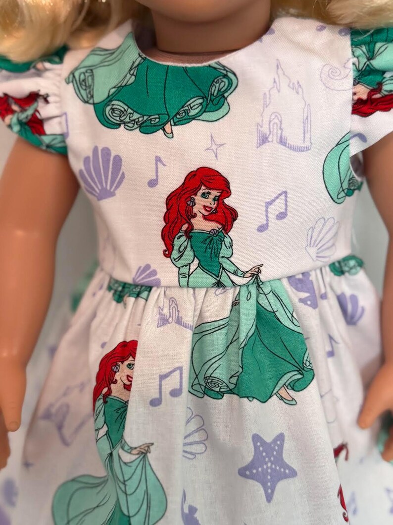 May include: A white doll dress featuring a repeating pattern of a red-haired princess in a green dress, musical notes, castles, seashells, and starfish. The dress has short sleeves and a gathered waist. The doll's blonde hair is visible.