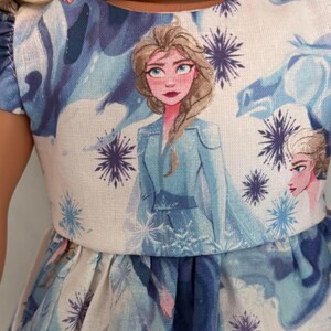 Puede incluir: Vestido de mu&ntilde;eca con un estampado de un personaje de la pel&iacute;cula animada "Frozen". El vestido tiene un corpi&ntilde;o blanco con un retrato del personaje y patrones de copos de nieve y hielo azules y morados. La falda es azul con un patr&oacute;n similar.