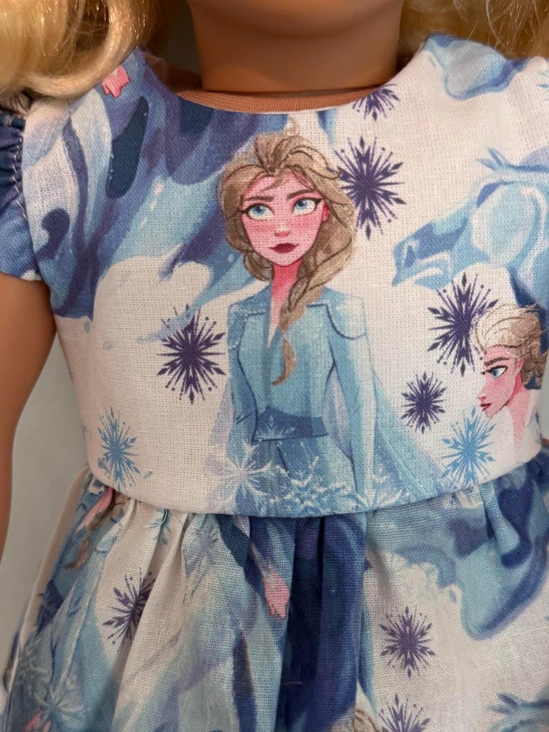 Puede incluir: Vestido de mu&ntilde;eca con un estampado de un personaje de la pel&iacute;cula animada "Frozen". El vestido tiene un corpi&ntilde;o blanco con un retrato del personaje y patrones de copos de nieve y hielo azules y morados. La falda es azul con un patr&oacute;n similar.